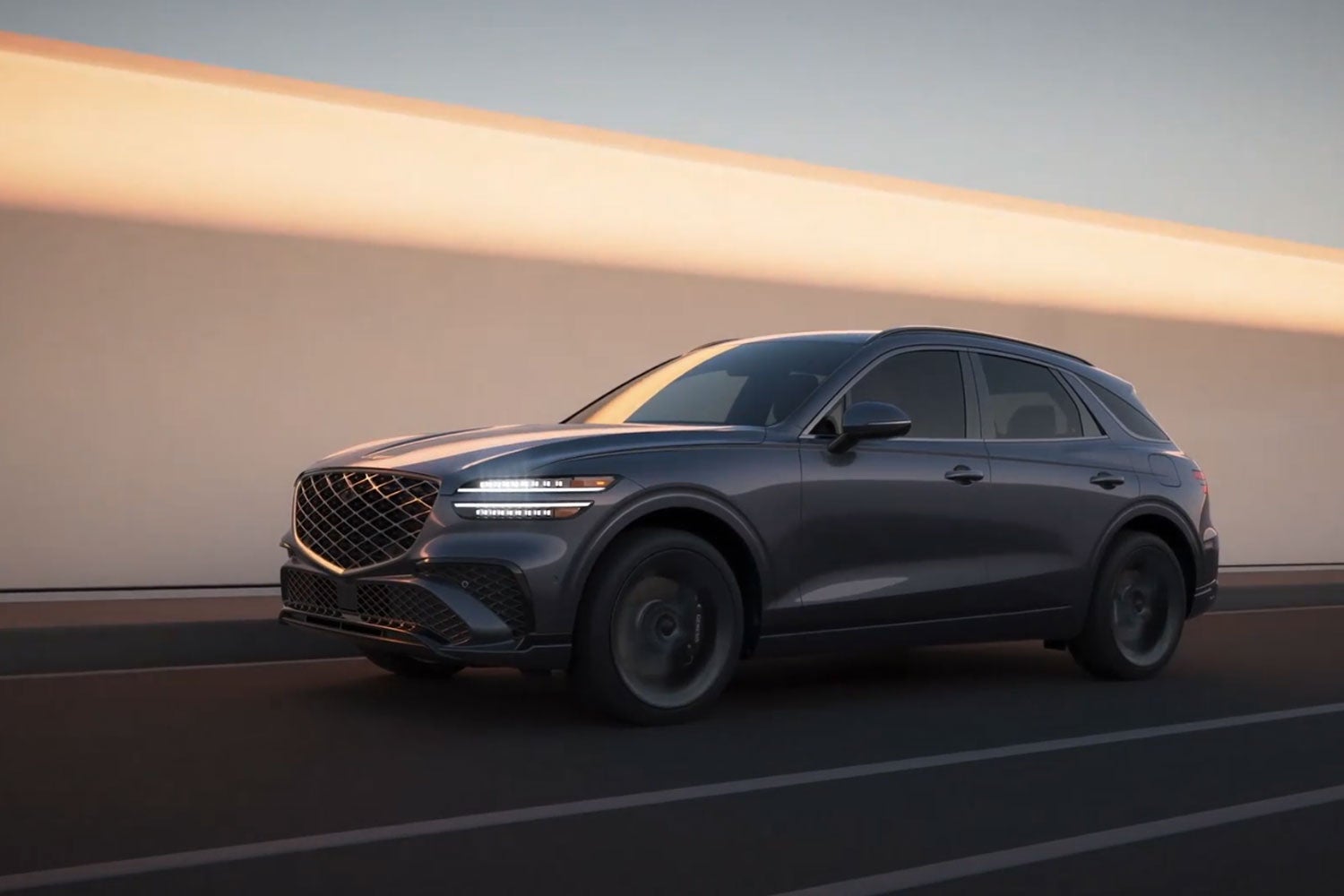 Dark grey Genesis GV70 SUV with headlights on, moving left to right against a sunlit beige wall.