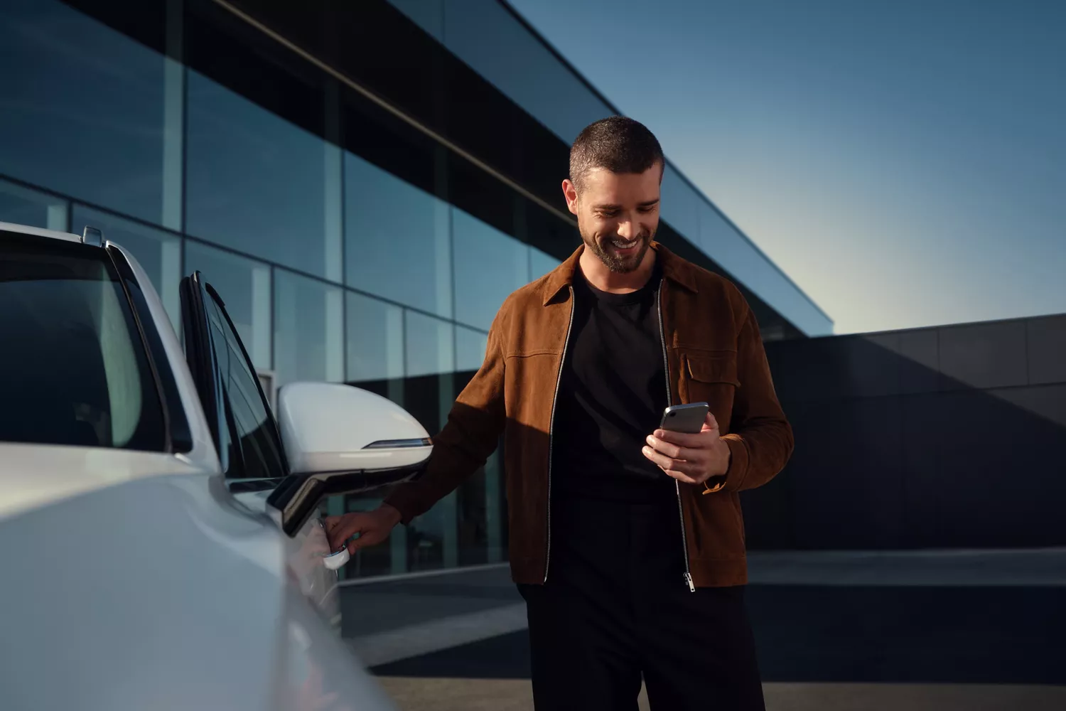 A smiling man in a brown jacket stands by a white car, holding a smartphone and reaching for the car door.